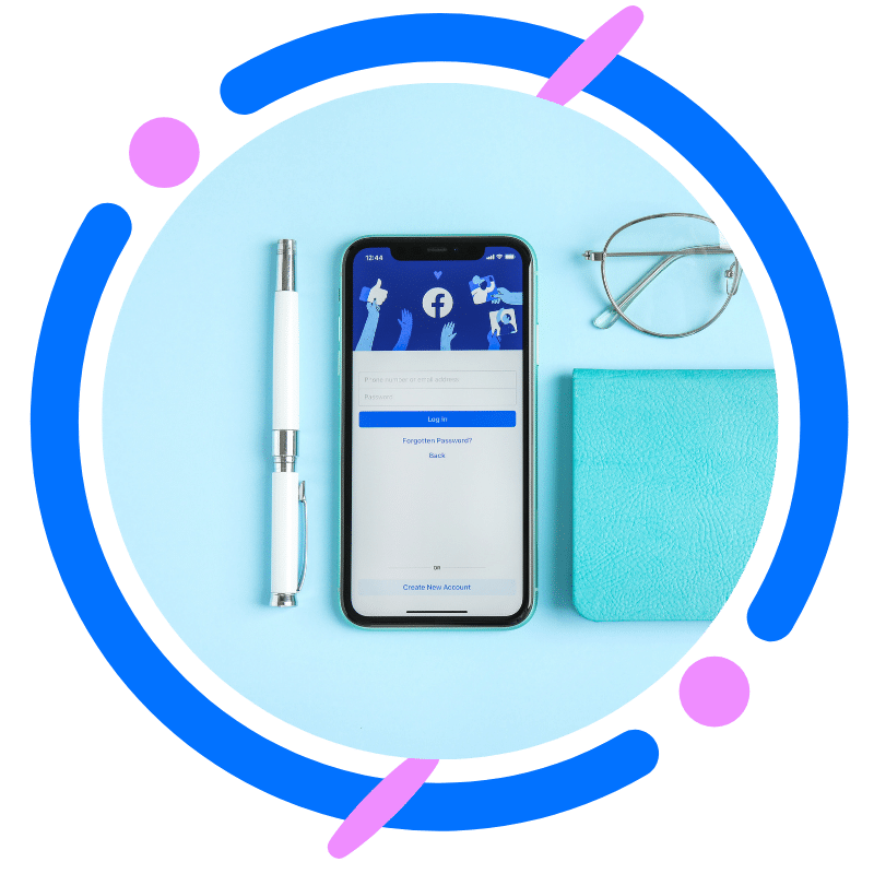 A smartphone displaying the Facebook login page on a blue background with a pen, glasses, and a teal notebook.